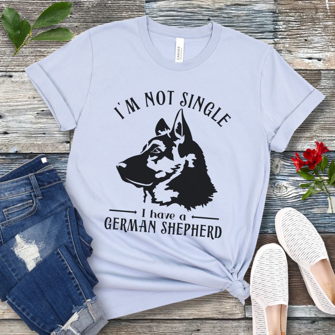 Light blue t-shirt with a German Shepherd graphic and 'I'm Not Single, I have a German Shepherd' text, presented on a wooden surface with jeans and shoes.