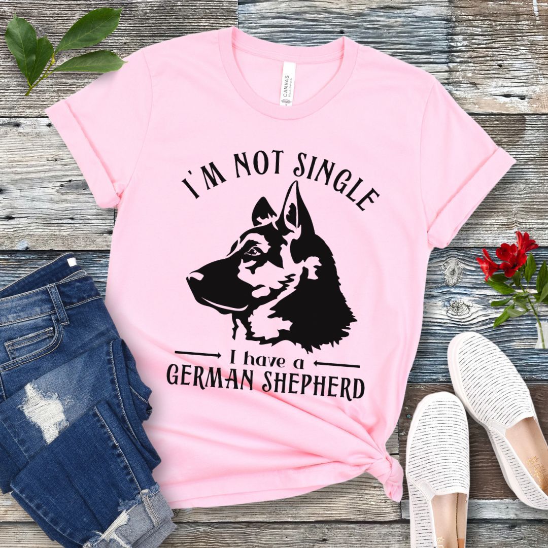 Pink t-shirt with a German Shepherd graphic and 'I'm Not Single, I have a German Shepherd' text, presented on a wooden surface with jeans and shoes.