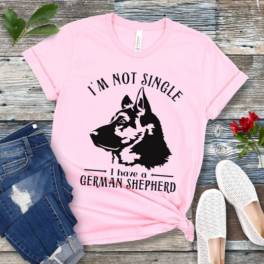 Pink t-shirt with a German Shepherd graphic and 'I'm Not Single, I have a German Shepherd' text, presented on a wooden surface with jeans and shoes.