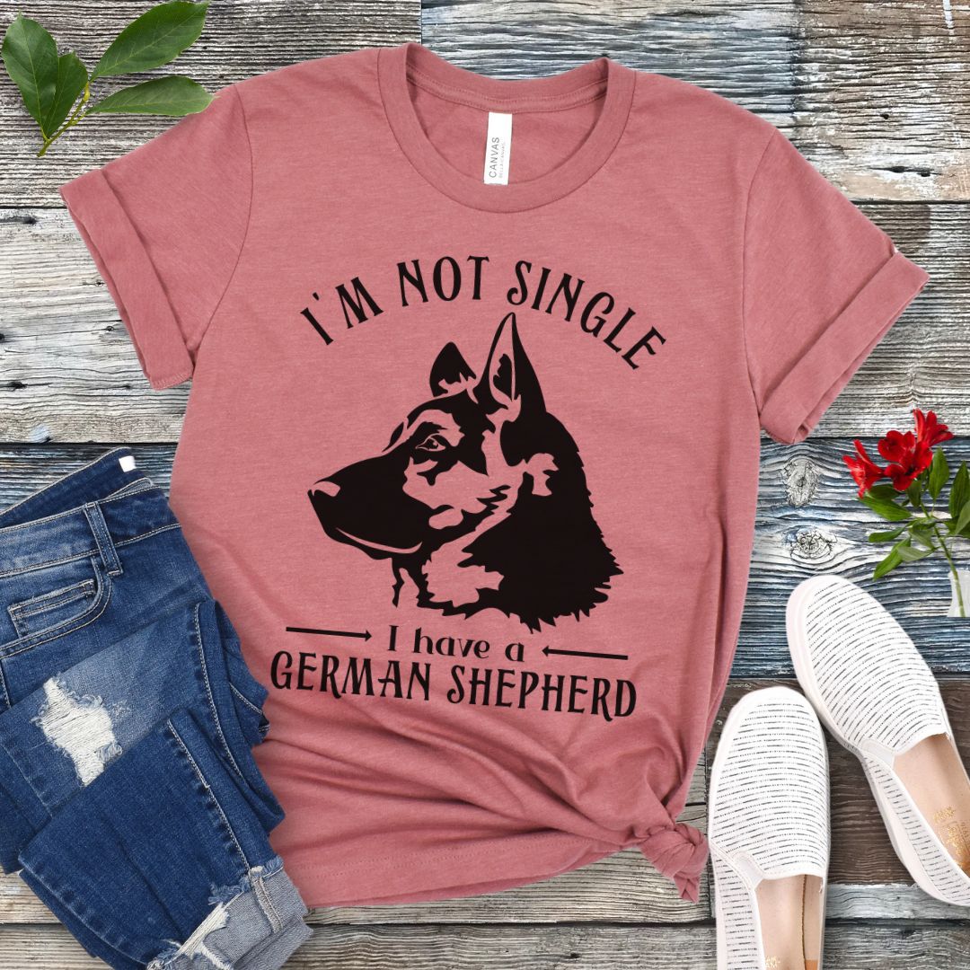 Mauve t-shirt with a German Shepherd graphic and 'I'm Not Single, I have a German Shepherd' text, presented on a wooden surface with jeans and shoes.