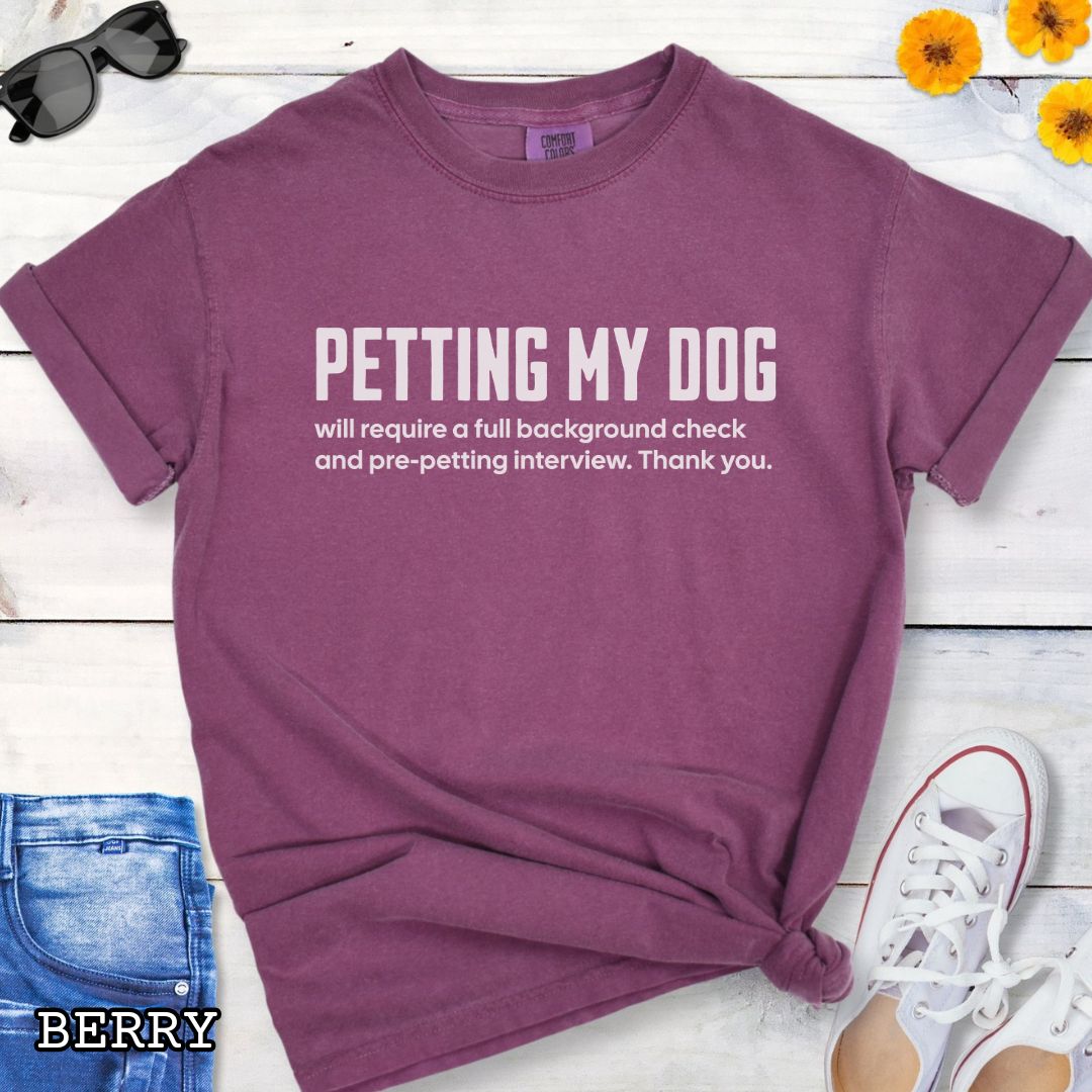 Berry Comfort Colors t-shirt for dog lovers with humorous text 'Petting My Dog will require a full background check and pre-petting interview. Thank you.', shown on a wooden surface with shoes and jeans.