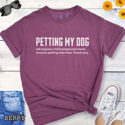 Berry Comfort Colors t-shirt for dog lovers with humorous text 'Petting My Dog will require a full background check and pre-petting interview. Thank you.', shown on a wooden surface with shoes and jeans.