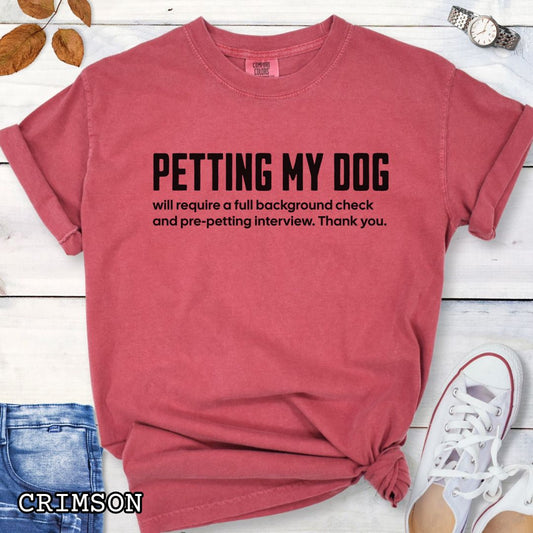 Red Comfort Colors t-shirt for dog lovers with humorous text 'Petting My Dog will require a full background check and pre-petting interview. Thank you.', shown on a wooden surface with shoes and jeans.