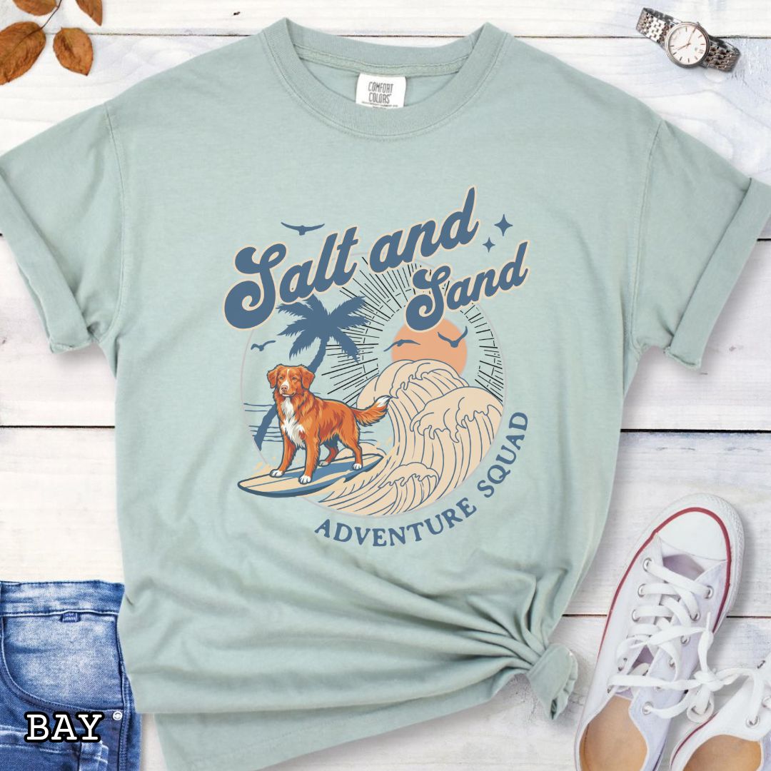Bay Comfort Colors t-shirt for Nova Scotia Duck Tolling Retriever lovers with beach-themed graphic of a surfing Toller dog, waves and sunset, and the text 'Salt and Sand Adventure Squad' in retro lettering, shown on a wooden surface with accessories.