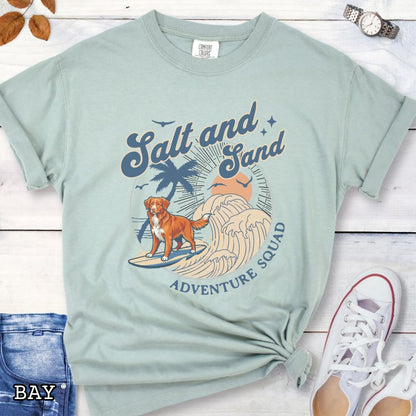 Bay Comfort Colors t-shirt for Nova Scotia Duck Tolling Retriever lovers with beach-themed graphic of a surfing Toller dog, waves and sunset, and the text 'Salt and Sand Adventure Squad' in retro lettering, shown on a wooden surface with accessories.