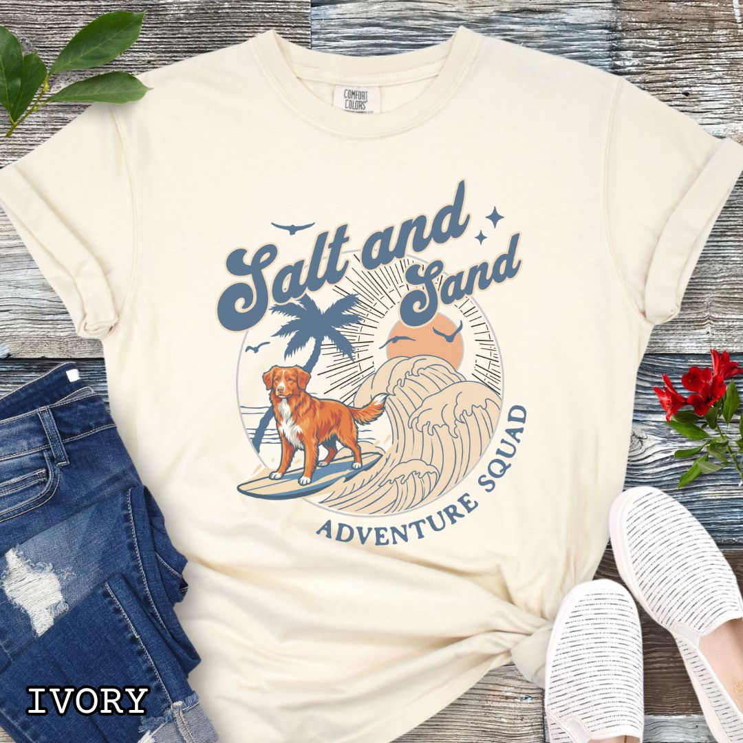 Beige Comfort Colors t-shirt for Nova Scotia Duck Tolling Retriever lovers with beach-themed graphic of a surfing Toller dog, waves and sunset, and the text 'Salt and Sand Adventure Squad' in retro lettering, shown on a wooden surface with accessories.