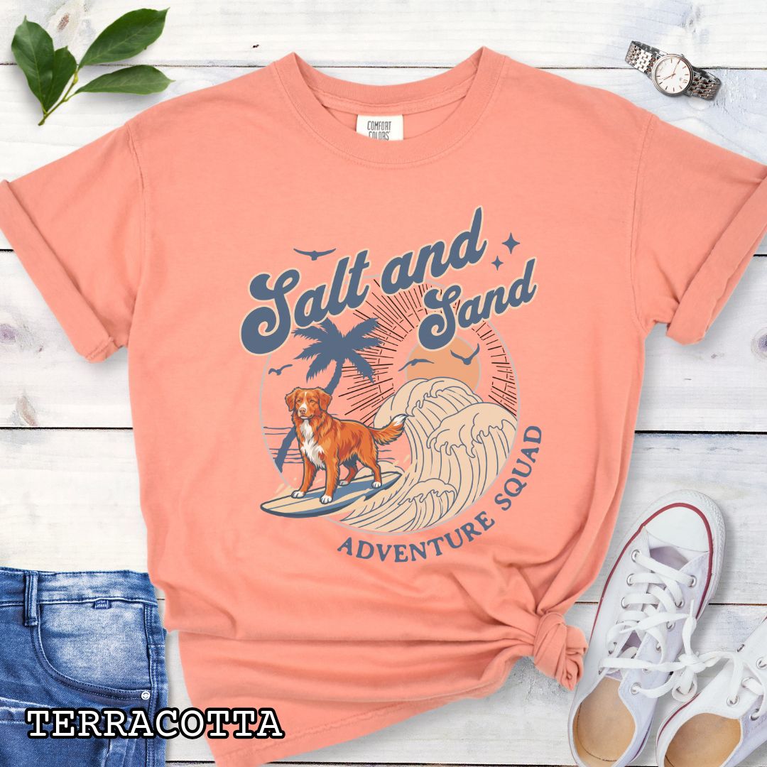Orange Comfort Colors t-shirt for Nova Scotia Duck Tolling Retriever lovers with beach-themed graphic of a surfing Toller dog, waves and sunset, and the text 'Salt and Sand Adventure Squad' in retro lettering, shown on a wooden surface with accessories.