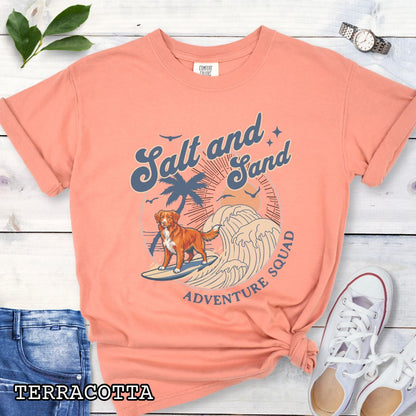 Orange Comfort Colors t-shirt for Nova Scotia Duck Tolling Retriever lovers with beach-themed graphic of a surfing Toller dog, waves and sunset, and the text 'Salt and Sand Adventure Squad' in retro lettering, shown on a wooden surface with accessories.