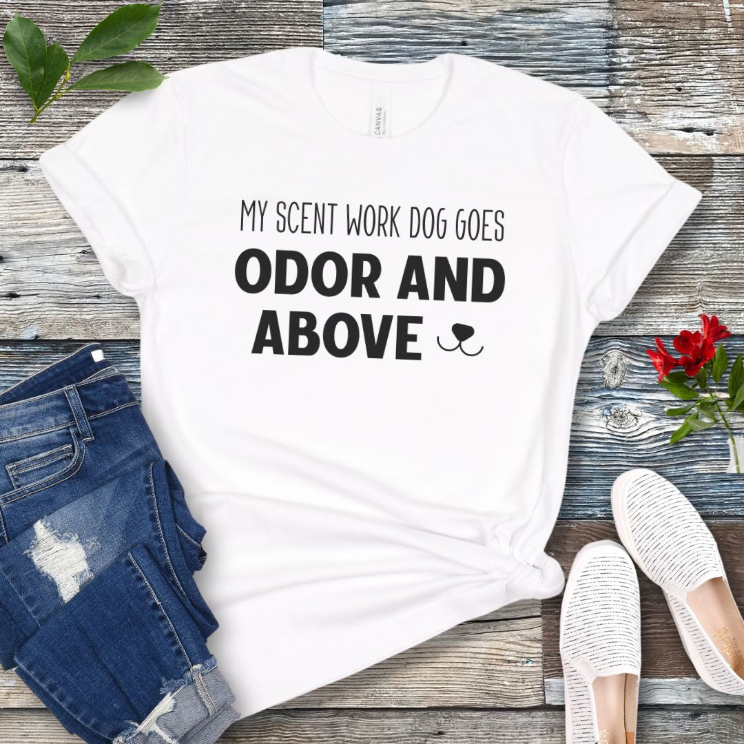 White dog sports t-shirt with text 'My scent work dog goes odor and above' on a wooden surface with jeans and shoes.