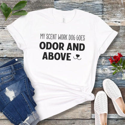 White dog sports t-shirt with text 'My scent work dog goes odor and above' on a wooden surface with jeans and shoes.