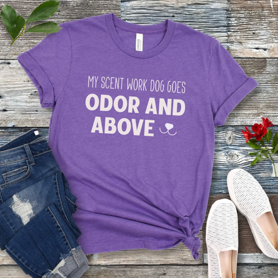 Purple dog sports t-shirt with text 'My scent work dog goes odor and above' on a wooden surface with jeans and shoes.