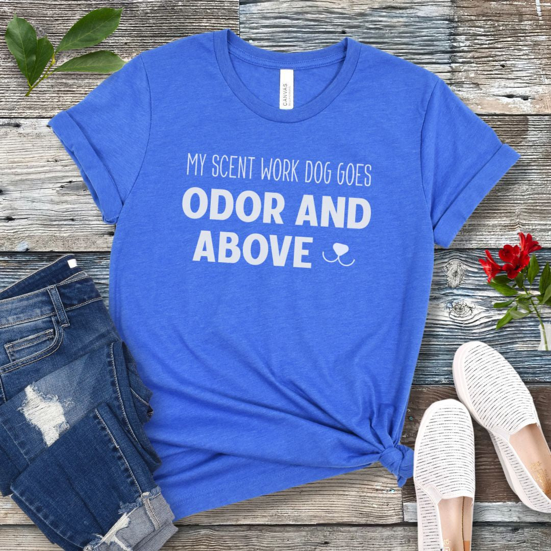 Blue dog sports t-shirt with text 'My scent work dog goes odor and above' on a wooden surface with jeans and shoes.
