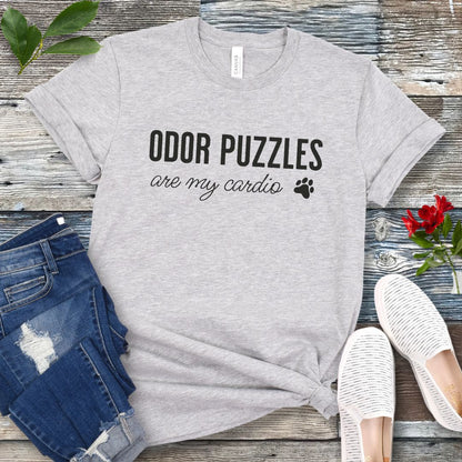 Grey canine scent work t-shirt with 'Odor Puzzles are my cardio' text on a wooden surface with jeans and shoes.