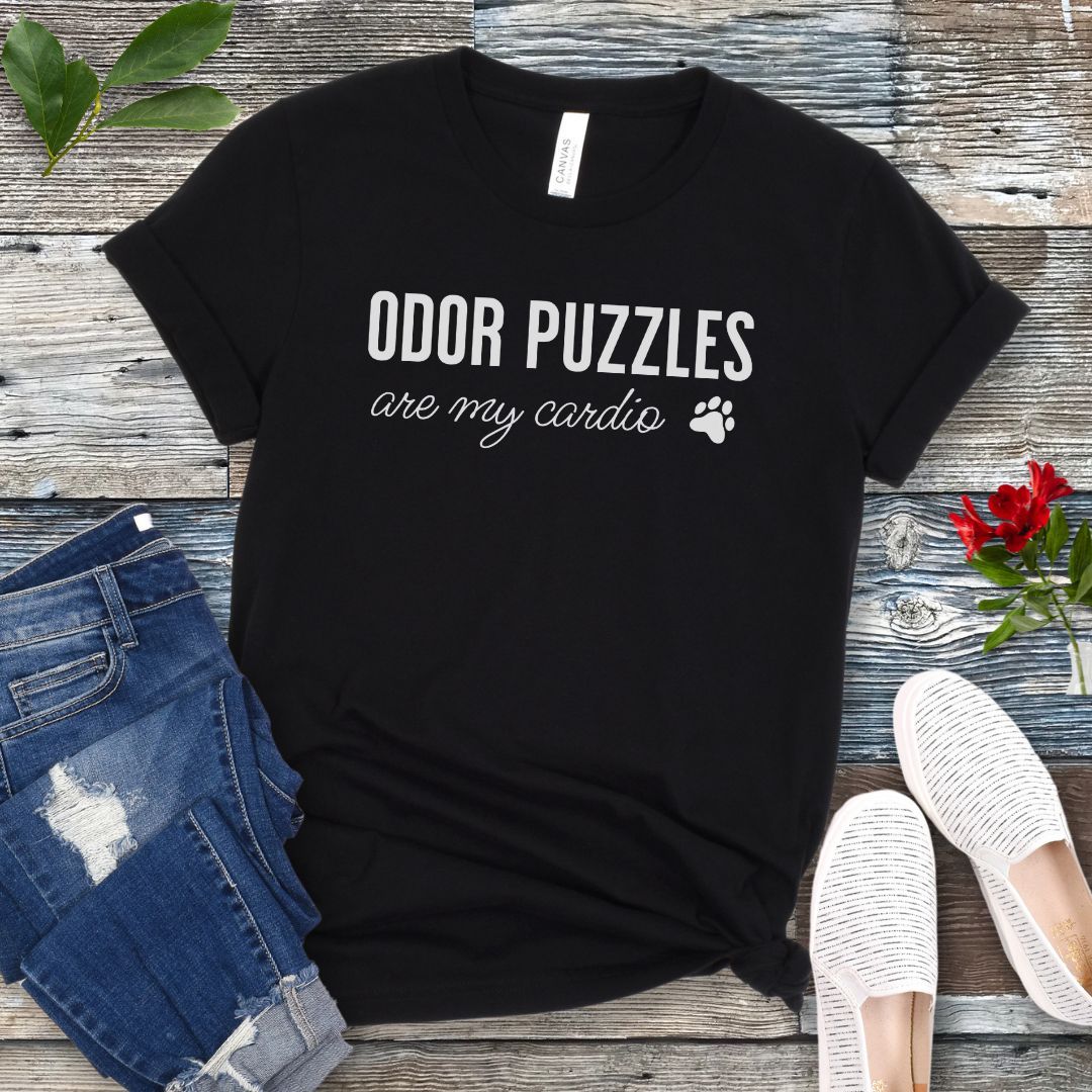 Black nosework for dogs t-shirt with 'Odor Puzzles are my cardio' text on a wooden surface with jeans and shoes.