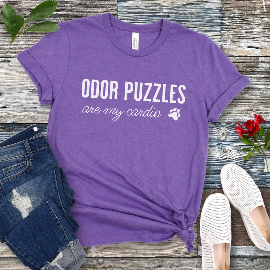 Purple canine scent detection t-shirt with 'Odor Puzzles are my cardio' text on a wooden surface with jeans and shoes.