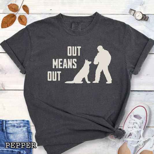 Dark gray Comfort Colors bitework t-shirt with a graphic of a K9 decoy and alertly sitting shepherd dog with text 'OUT MEANS OUT', shown with accessories on a wooden surface.