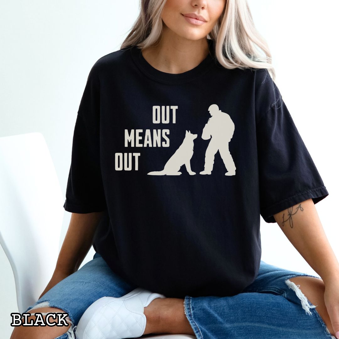 Black Comfort Colors bitework t-shirt with a graphic of a K9 decoy and alertly sitting shepherd dog with text 'OUT MEANS OUT', shown with accessories on a wooden surface.