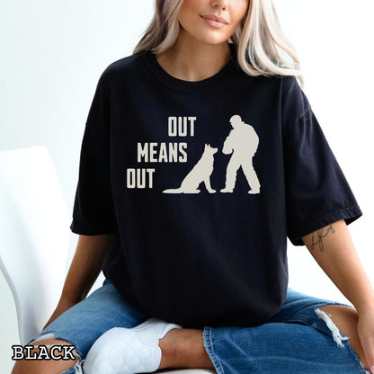 Black Comfort Colors bitework t-shirt with a graphic of a K9 decoy and alertly sitting shepherd dog with text 'OUT MEANS OUT', shown with accessories on a wooden surface.