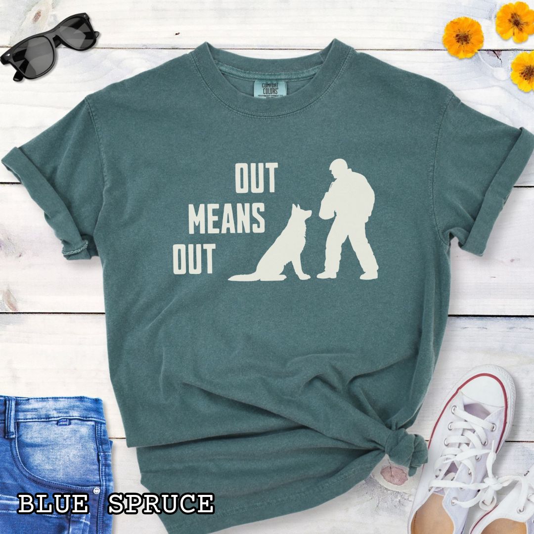 Green Comfort Colors bitework t-shirt with a graphic of a K9 decoy and alertly sitting shepherd dog with text 'OUT MEANS OUT', shown with accessories on a wooden surface.