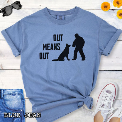 Blue Comfort Colors bitework t-shirt with a graphic of a K9 decoy and alertly sitting shepherd dog with text 'OUT MEANS OUT', shown with accessories on a wooden surface.