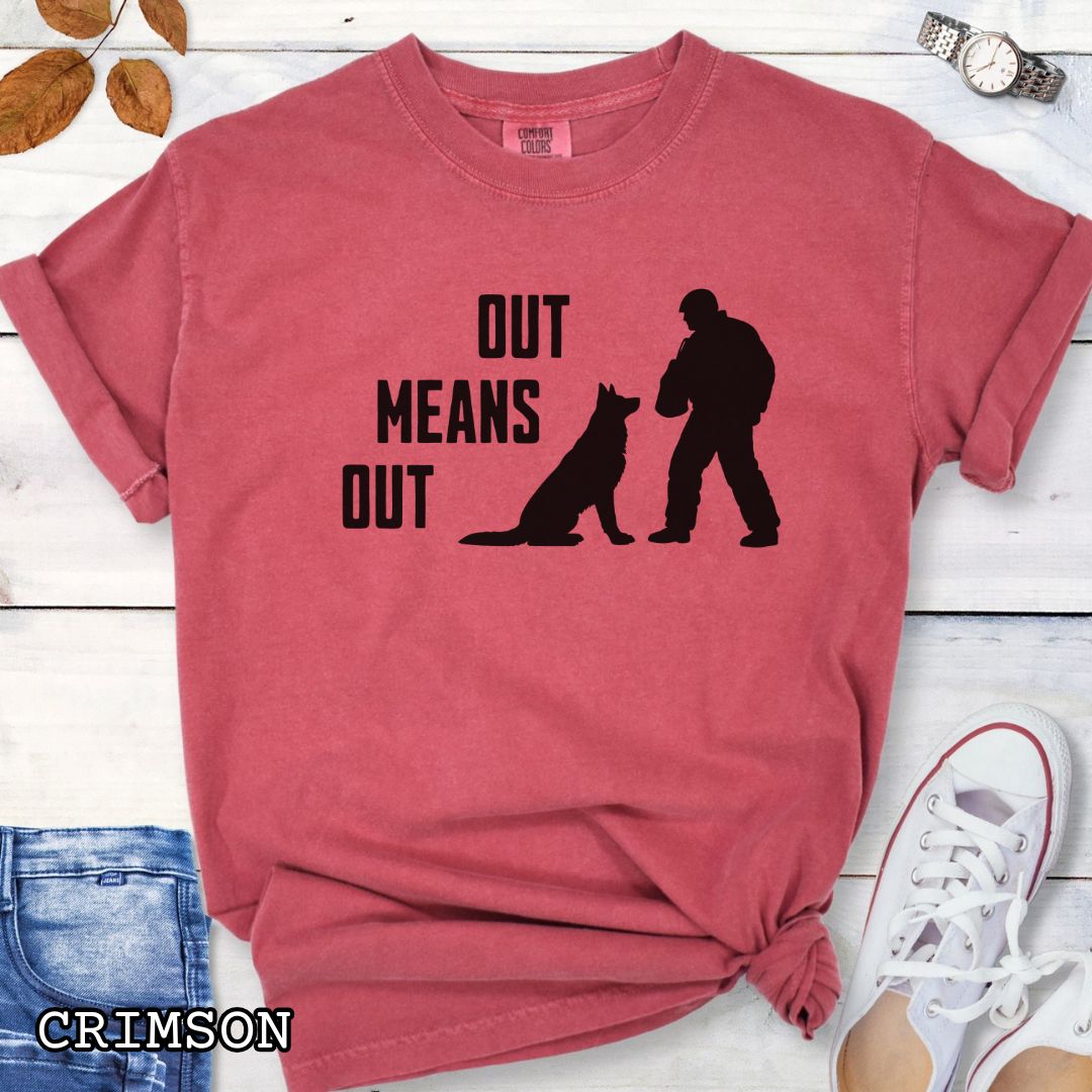 Red Comfort Colors bitework t-shirt with a graphic of a K9 decoy and alertly sitting shepherd dog with text 'OUT MEANS OUT', shown with accessories on a wooden surface.