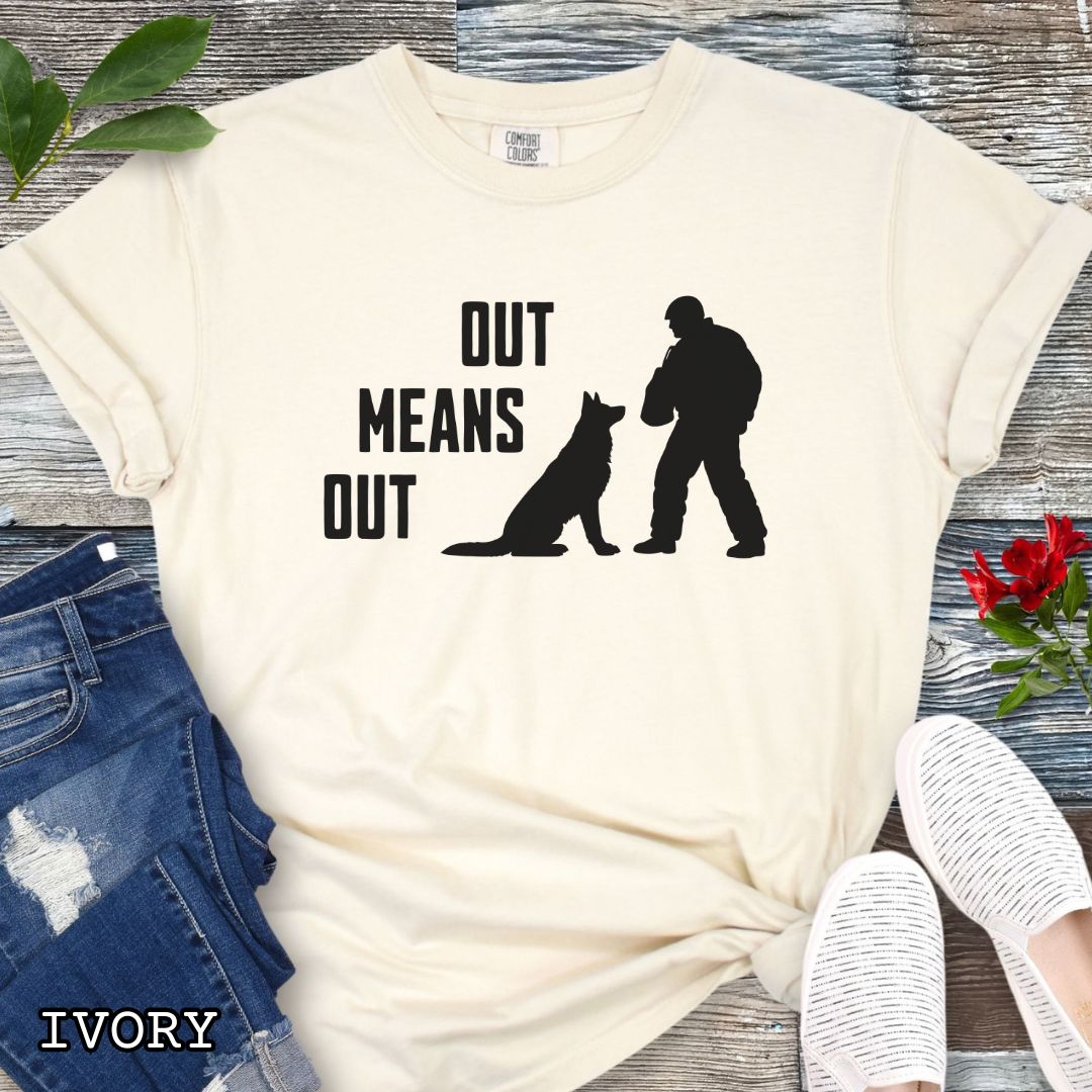 Ivory Comfort Colors bitework t-shirt with a graphic of a K9 decoy and alertly sitting shepherd dog with text 'OUT MEANS OUT', shown with accessories on a wooden surface.