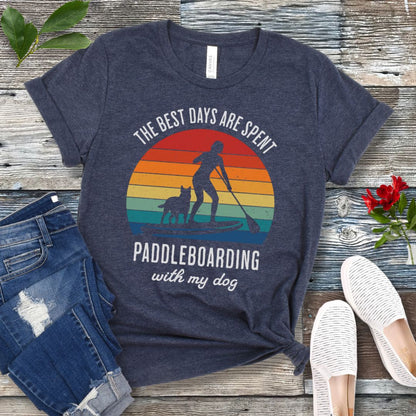 A heather navy t-shirt for dog lovers with a graphic print of a vintage sunset, a person paddleboarding with a dog, and the text 'THE BEST DAYS ARE SPENT PADDLEBOARDING WITH MY DOG'. The shirt is presented on a wooden surface with a pair of jeans and white shoes in the background.