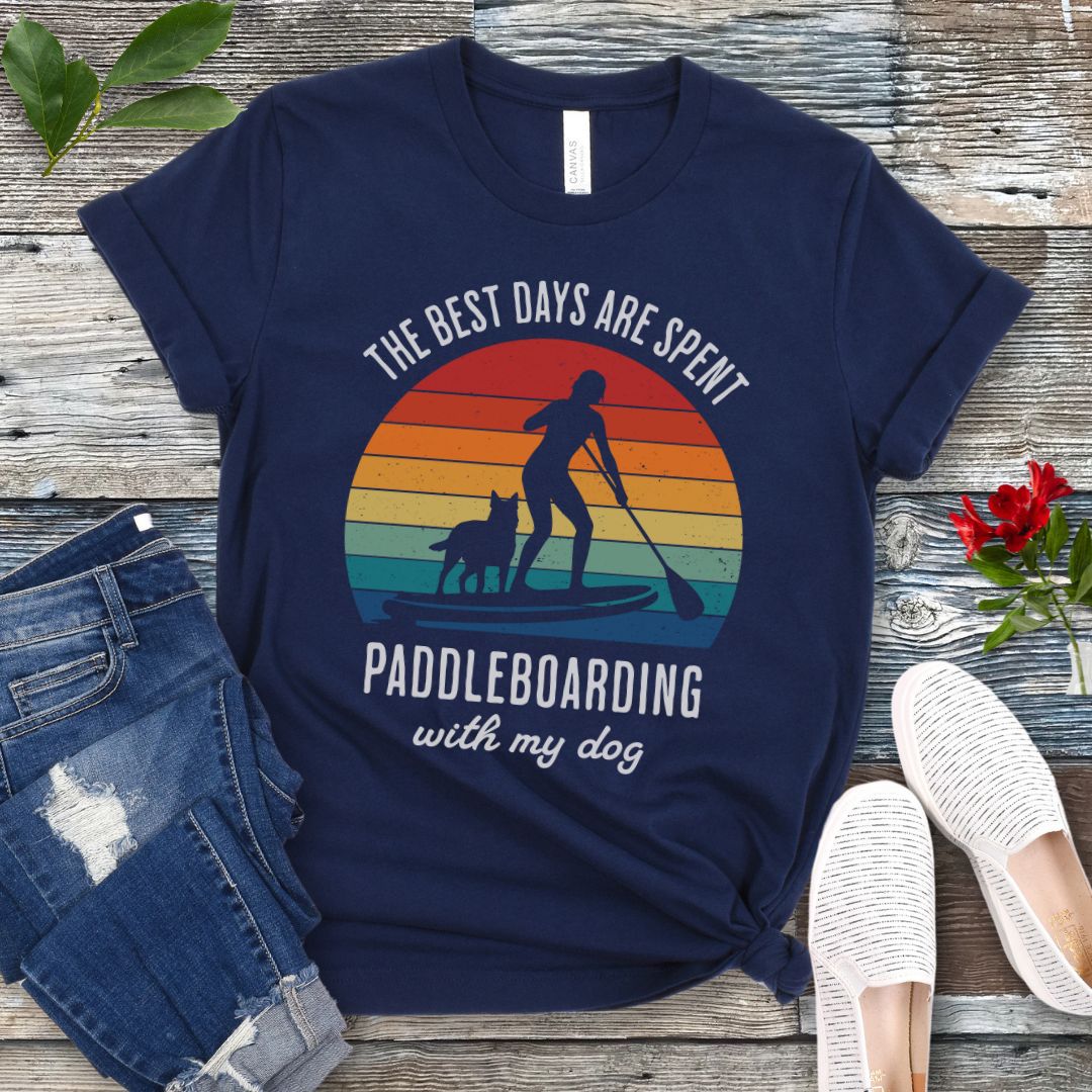 A navy blue t-shirt for dog lovers with a graphic print of a vintage sunset, a person paddleboarding with a dog, and the text 'THE BEST DAYS ARE SPENT PADDLEBOARDING WITH MY DOG'. The shirt is presented on a wooden surface with a pair of jeans and white shoes in the background.