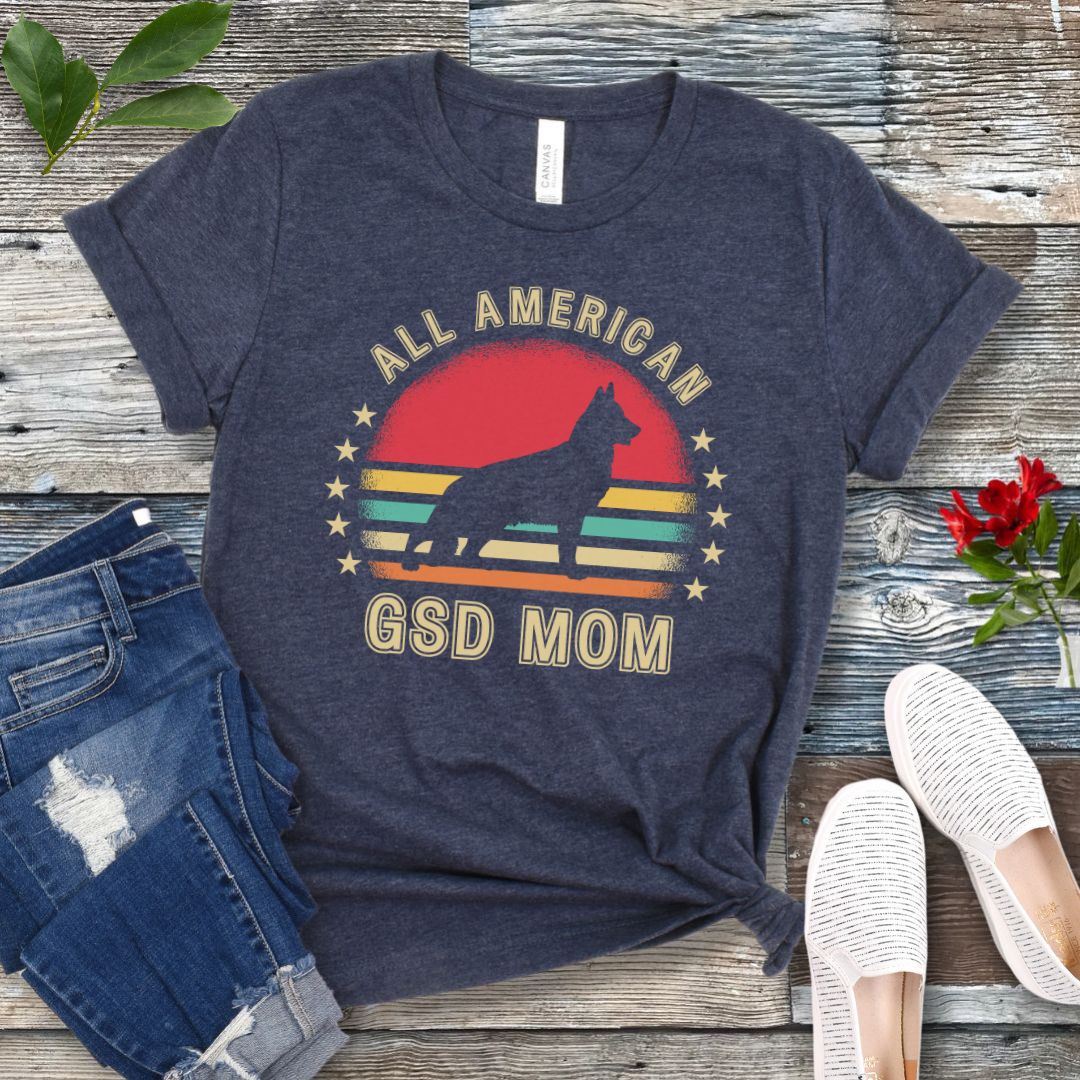 Heather navy t-shirt with colorful vintage sunset, stars, and the text 'All American GSD Mom', presented on a wooden surface with jeans and shoes.