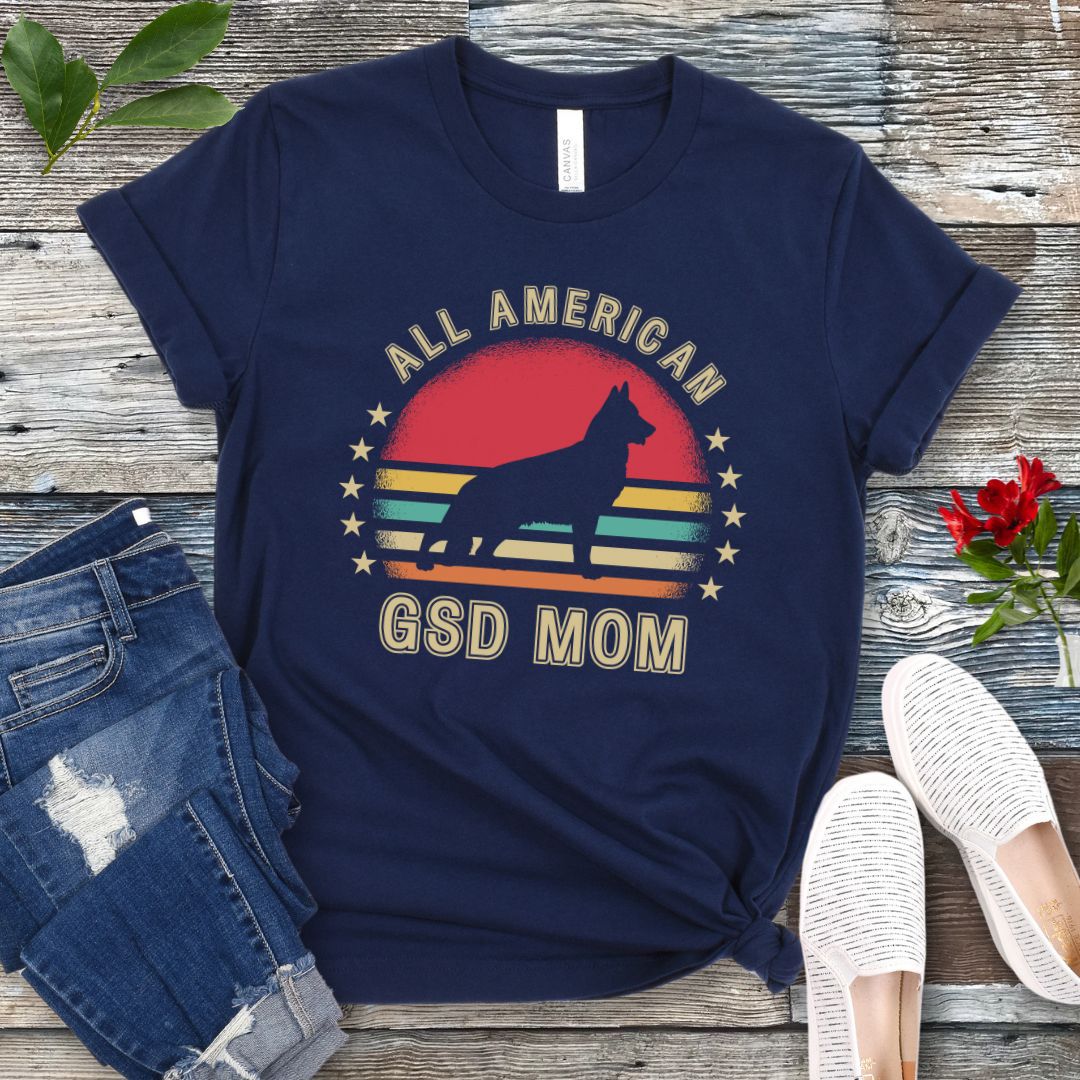 Navy blue t-shirt with colorful vintage sunset, stars, and the text 'All American GSD Mom', presented on a wooden surface with jeans and shoes.