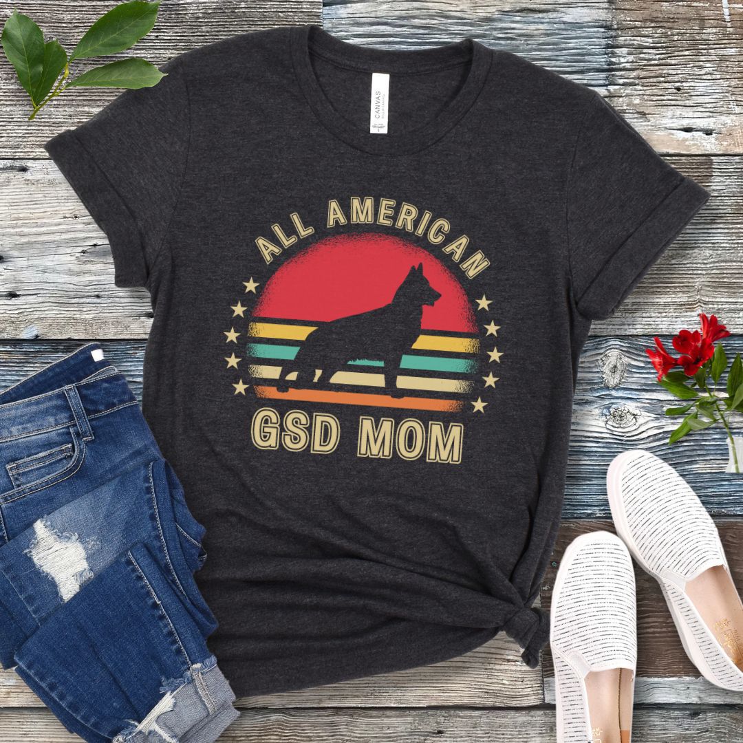 Dark grey t-shirt with colorful vintage sunset, stars, and the text 'All American GSD Mom', presented on a wooden surface with jeans and shoes.