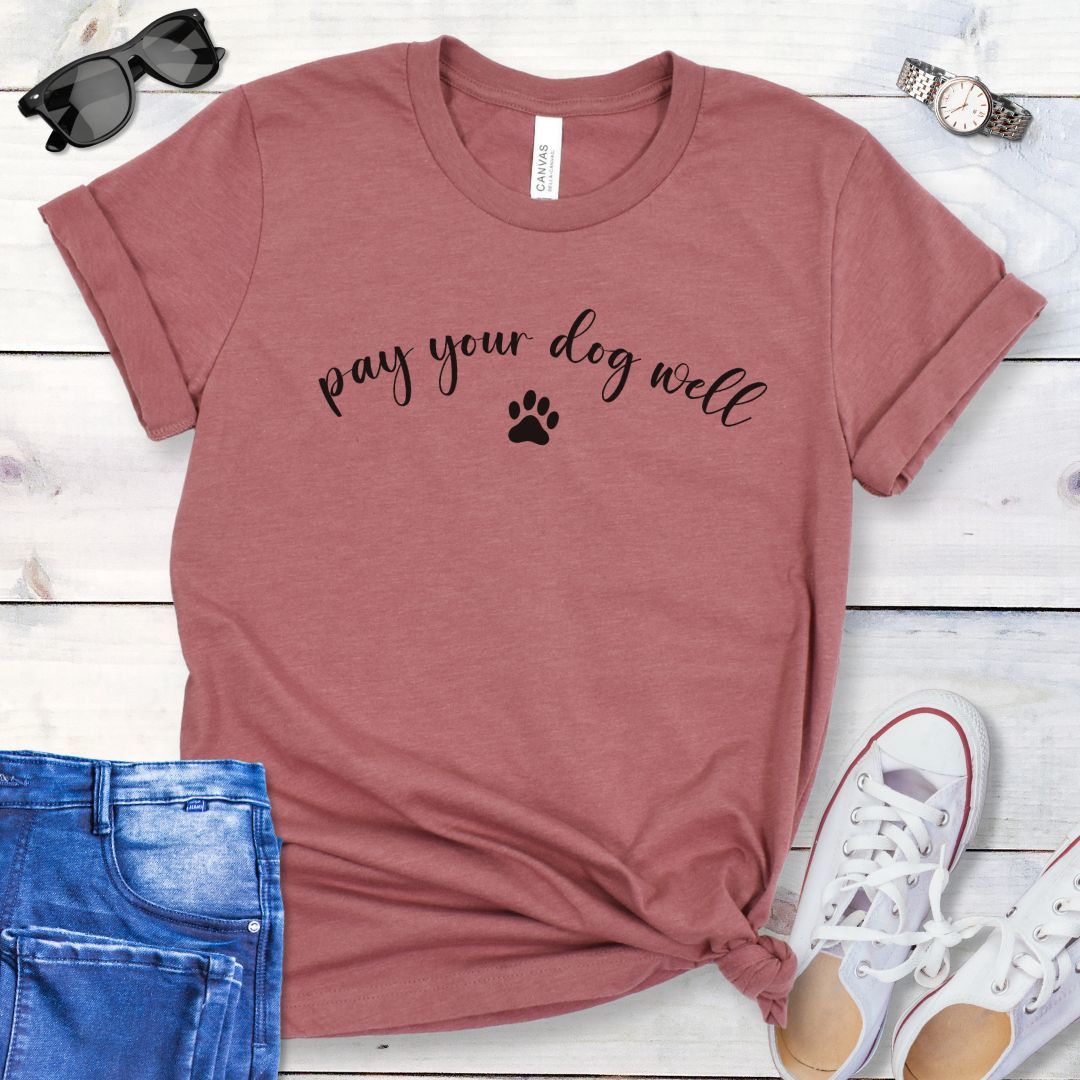 Mauve dog training t-shirt with 'pay your dog well' text in script and paw print, shown on a wooden surface with jeans and shoes.