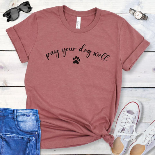 Mauve dog training t-shirt with 'pay your dog well' text in script and paw print, shown on a wooden surface with jeans and shoes.