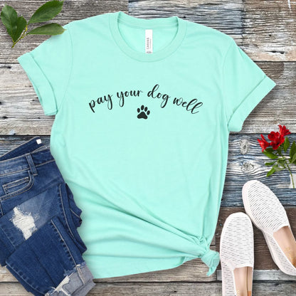 Mint green dog training t-shirt with 'pay your dog well' text and paw print on a wooden surface with jeans and shoes.