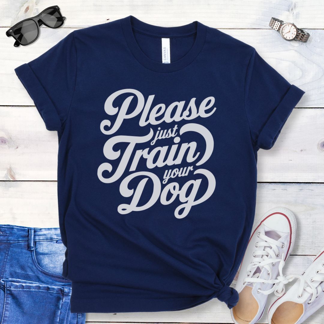 A navy t-shirt with the text 'Please Just Train your Dog' printed in a playful, bold font. The t-shirt is presented in a casual style, accompanied by jeans and white shoes, suggesting a casual setting.