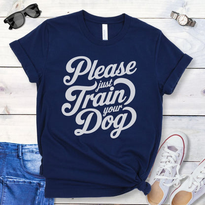 A navy t-shirt with the text 'Please Just Train your Dog' printed in a playful, bold font. The t-shirt is presented in a casual style, accompanied by jeans and white shoes, suggesting a casual setting.