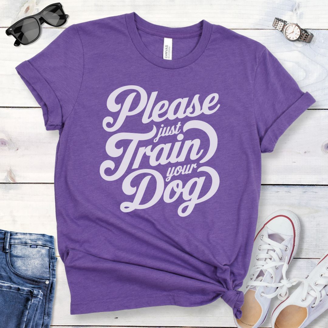 A purple t-shirt with the text 'Please Just Train your Dog' printed in a playful, bold font. The t-shirt is presented in a casual style, accompanied by jeans and white shoes, suggesting a casual setting.