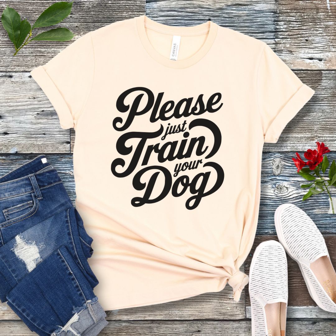 A beige-colored t-shirt with the text 'Please Just Train your Dog' printed in a playful, bold font. The t-shirt is presented in a casual style, accompanied by jeans and white shoes, suggesting a casual setting.