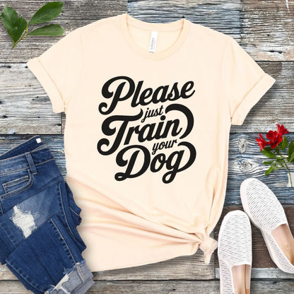 A beige-colored t-shirt with the text 'Please Just Train your Dog' printed in a playful, bold font. The t-shirt is presented in a casual style, accompanied by jeans and white shoes, suggesting a casual setting.