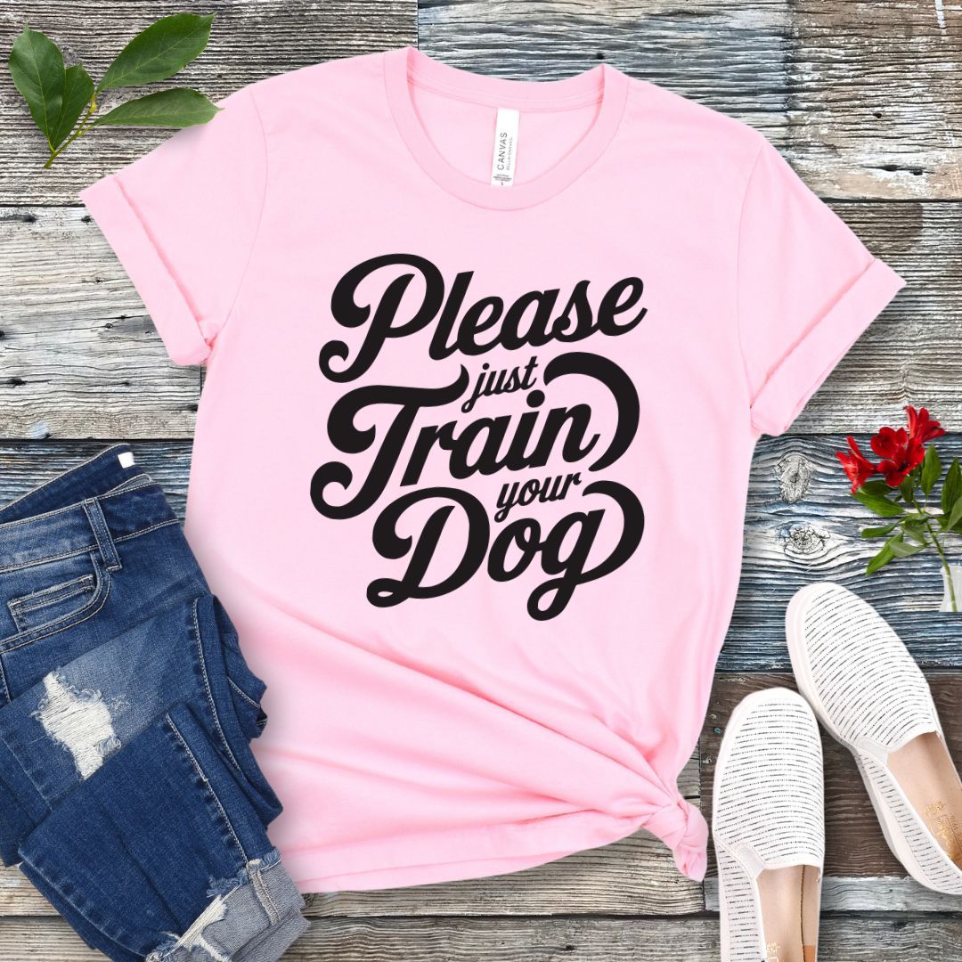 A pink t-shirt with the text 'Please Just Train your Dog' printed in a playful, bold font. The t-shirt is presented in a casual style, accompanied by jeans and white shoes, suggesting a casual setting.
