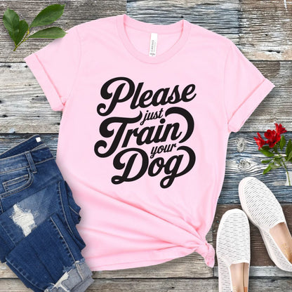 A pink t-shirt with the text 'Please Just Train your Dog' printed in a playful, bold font. The t-shirt is presented in a casual style, accompanied by jeans and white shoes, suggesting a casual setting.