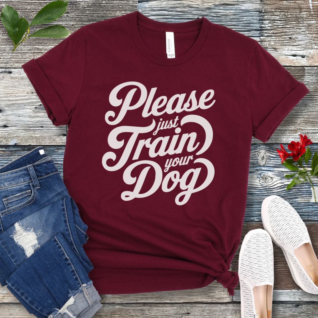 A maroon t-shirt with the text 'Please Just Train your Dog' printed in a playful, bold font. The t-shirt is presented in a casual style, accompanied by jeans and white shoes, suggesting a casual setting.