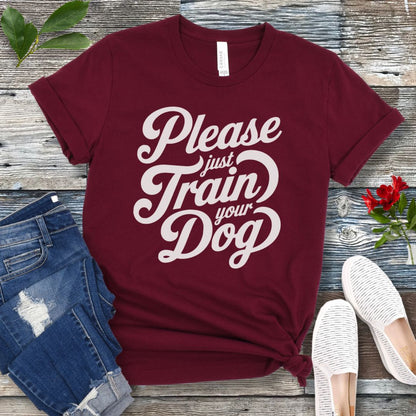 A maroon t-shirt with the text 'Please Just Train your Dog' printed in a playful, bold font. The t-shirt is presented in a casual style, accompanied by jeans and white shoes, suggesting a casual setting.