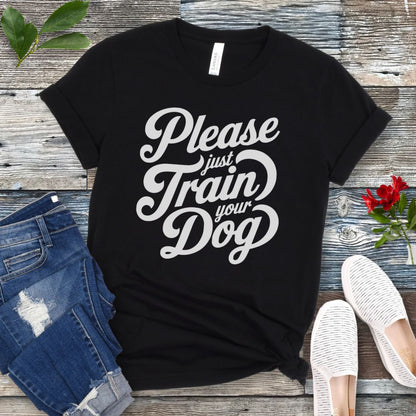 A black t-shirt with the text 'Please Just Train your Dog' printed in a playful, bold font. The t-shirt is presented in a casual style, accompanied by jeans and white shoes, suggesting a casual setting.