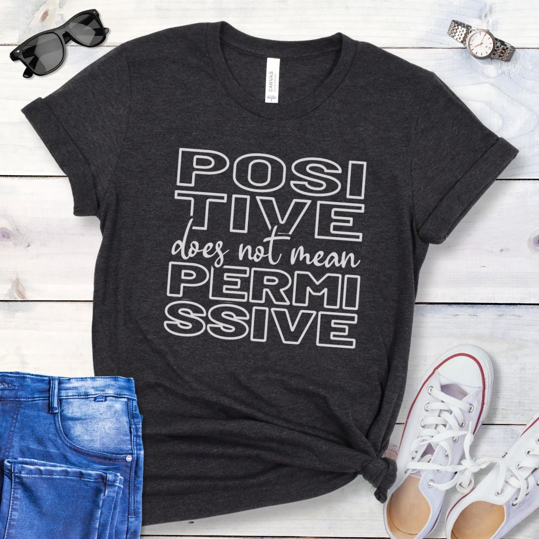 Dark grey dog training t-shirt with 'POSITIVE does not mean PERMISSIVE' typography text on a wooden surface with jeans and shoes.