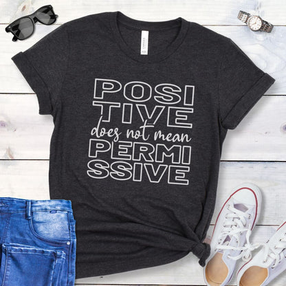 Dark grey dog training t-shirt with 'POSITIVE does not mean PERMISSIVE' typography text on a wooden surface with jeans and shoes.