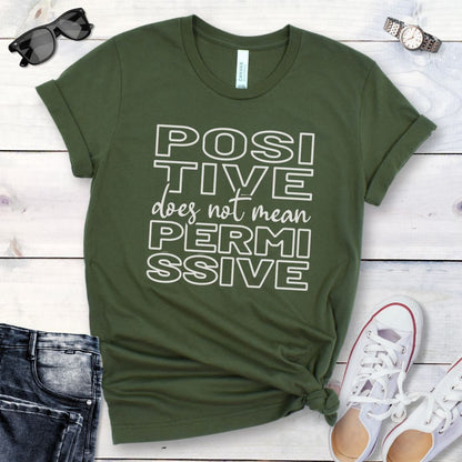 Military green dog training t-shirt with 'POSITIVE does not mean PERMISSIVE' typography text on a wooden surface with jeans and shoes.