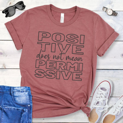 Mauve dog training t-shirt with 'POSITIVE does not mean PERMISSIVE' typography text on a wooden surface with jeans and shoes.