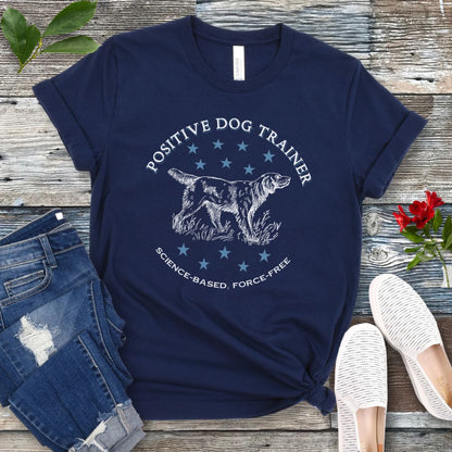 A navy blue t-shirt with a vintage dog illustration and text that reads 'POSITIVE DOG TRAINER SCIENCE-BASED FORCE-FREE', displayed on a wooden background with jeans and white shoes.