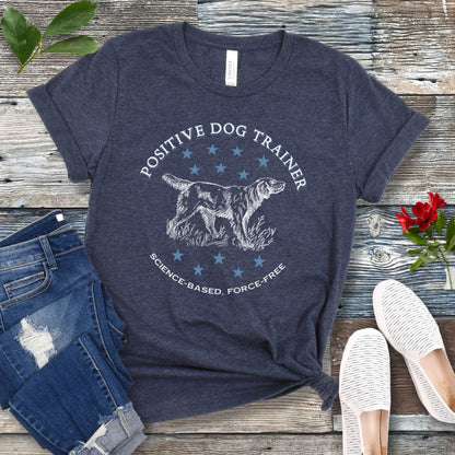 A heather navy t-shirt with a vintage dog illustration and text that reads 'POSITIVE DOG TRAINER SCIENCE-BASED FORCE-FREE', displayed on a wooden background with jeans and white shoes.